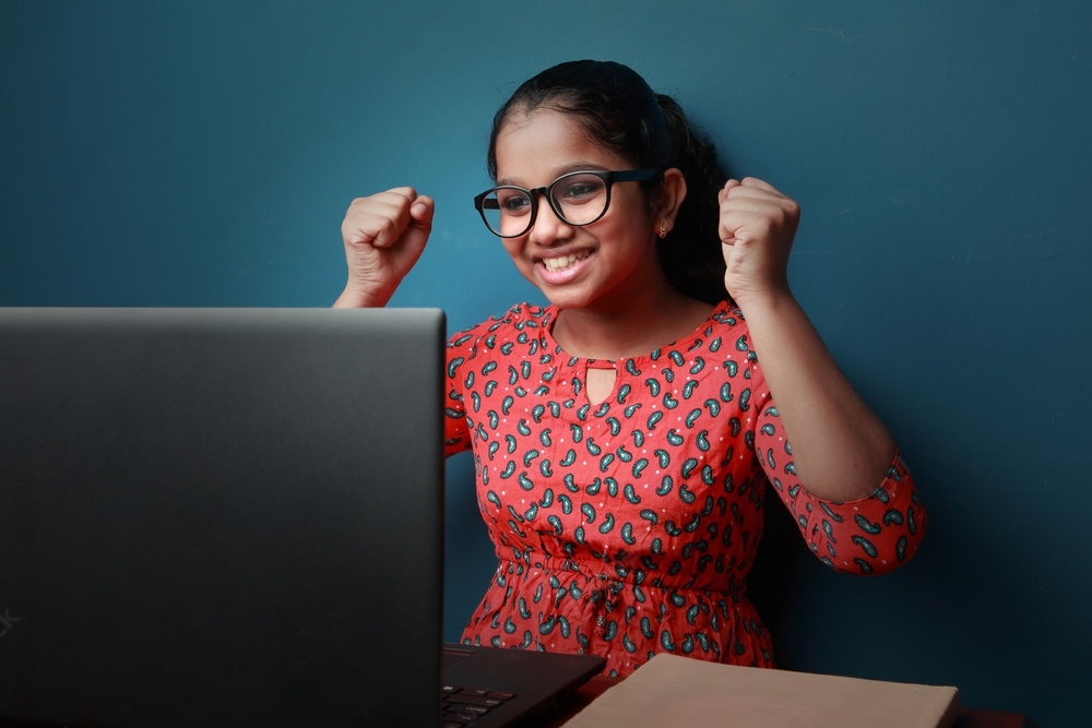A young girl wearing glasses and a red dress sits at a desk in front of a laptop in her home. She is smiling broadly and has her fists raised in a gesture of excitement or triumph. The background is a plain blue wall.