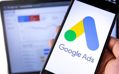 Hand holding smartphone displaying Google Ads logo with analytics dashboard in the background